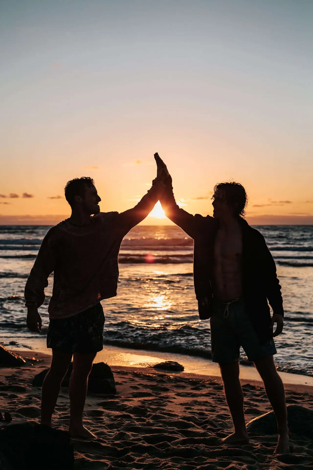 This photo shows two young man high fiving in the air and enjoying the sunset on the beach on their almost silhouette visual.